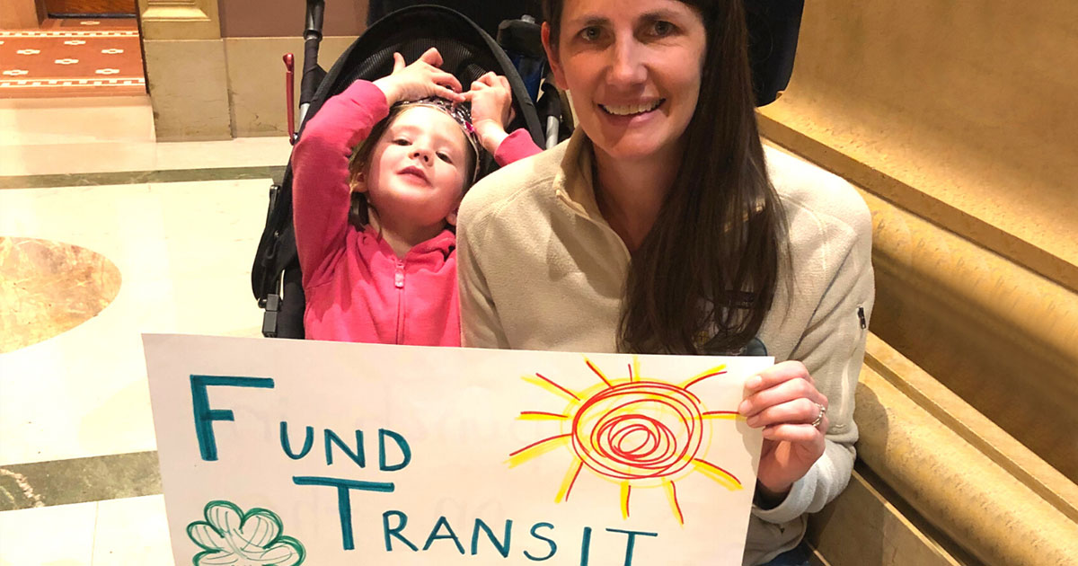 Person smiling with child in stroller, holding a sign that reads "Fund Transit"