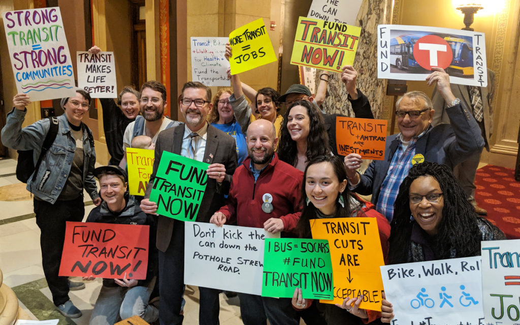 Transit advocates smile for a group photo at the MN State Capitol holding colorful signs with messages demanding investment in transit, bicycling, and walking.