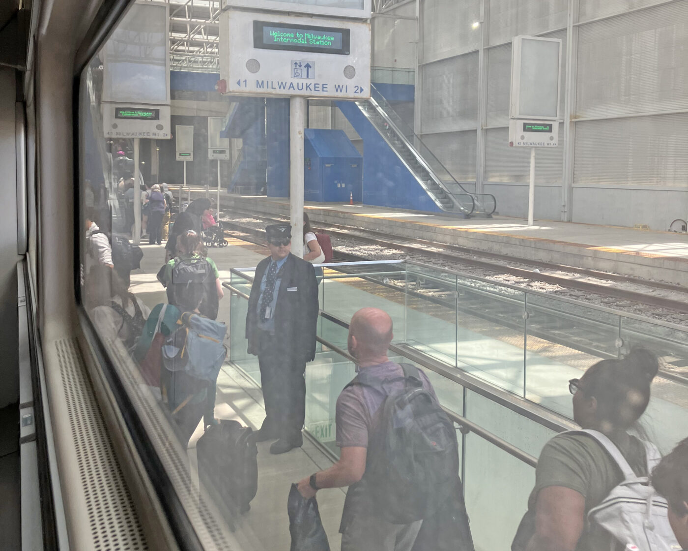 A train station in Milwaukee with a uniformed station attendant and a number of passengers walking alongside the train cars