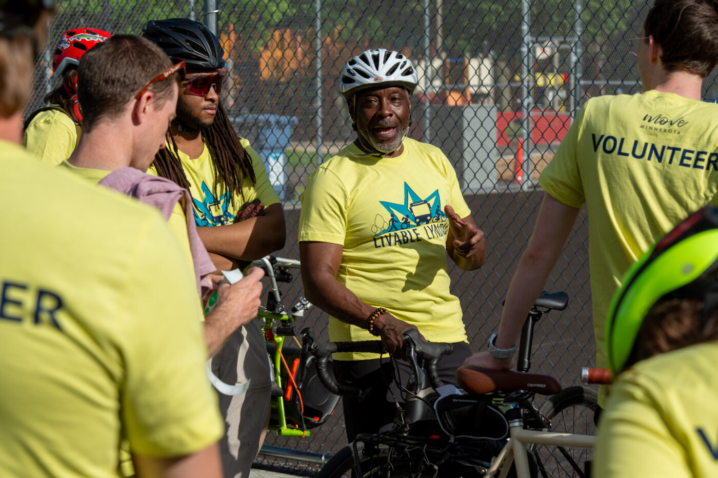 A Black man wearing a bright green Livable Lyndale t-shirt and a white bike helmet speaks to a gorup of volunteers 