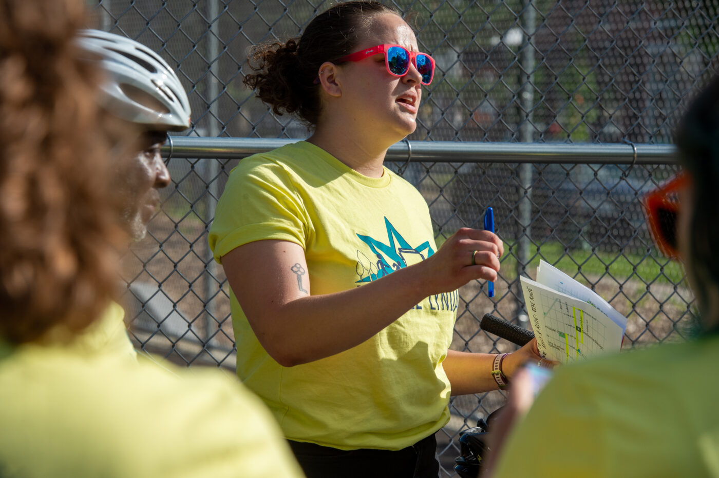 A white woman with curly hair wearing pink sunglasses and a bright green t-shirt talking to a group of volunteers at an outdoor rally