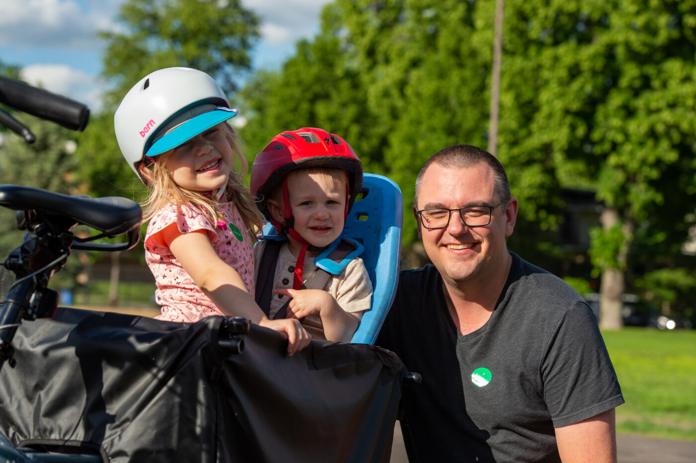 A man with a shaved head smiles next to two young children wearing bike helmets in a black bike trailer 