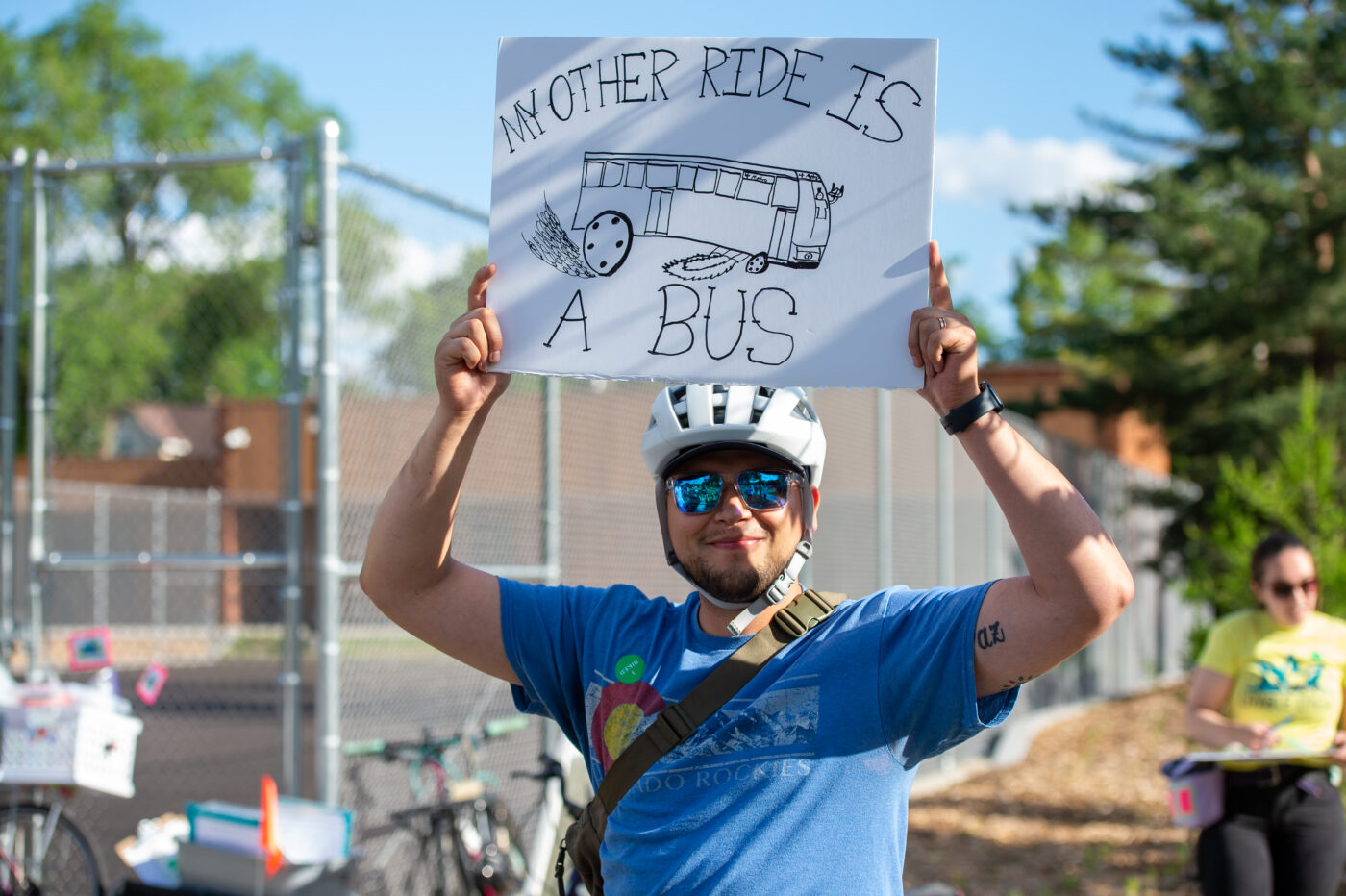 An advocate at a Livable Lyndale wearing a bike helmet and holding a sign that says "My Other Ride is A Bus"