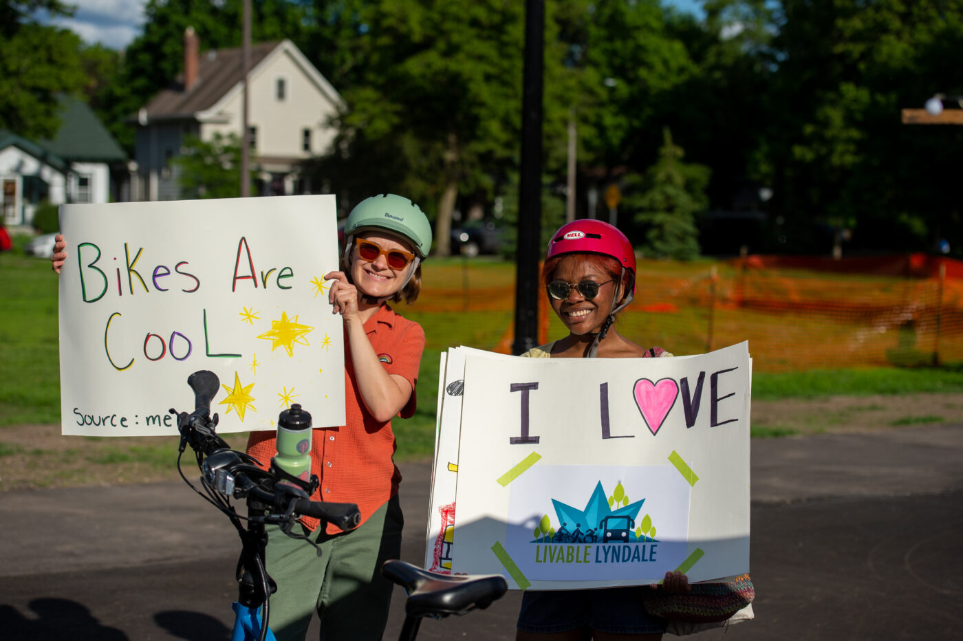 A young white bicyclist holding up a signs that reads "Bikes Are Cool" next to a young Black bicyclist with a sign that says "I Love Livable Lyndale"