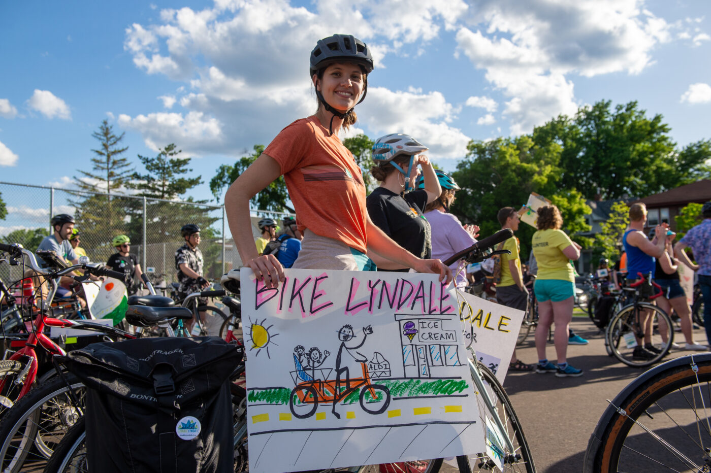 A young white woman wearing a bike helmet and orange t-shirt holds a handmade sign that says Bike Lyndale above a stick-figure family on a bicycle 