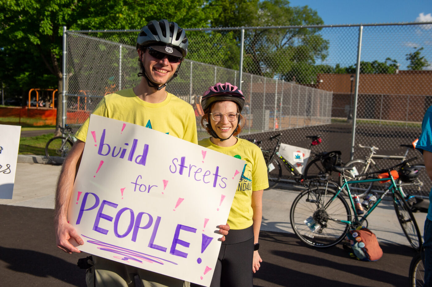 A young white couple wearing bike helmets and bright green t-shirts hold a sign that reads "Build Streets for PEOPLE"