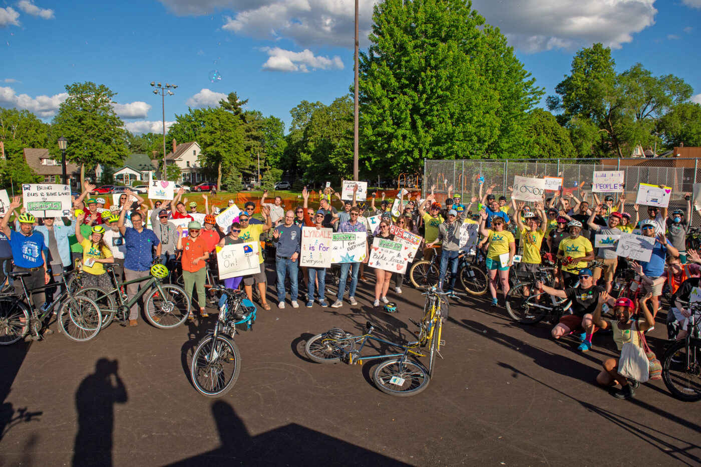 A group of close to 100 people holding handmade signs, many wearing bike helmets and raising their arms at to signal joy and energy for a Livable Lyndale