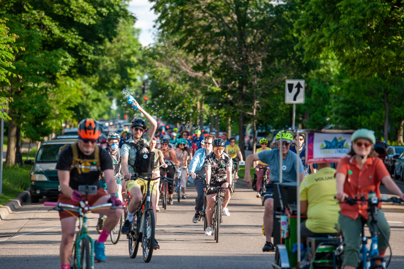 A vibrant group of bicyclists on a tree-lined street, with one rider blowing bubbles above his head