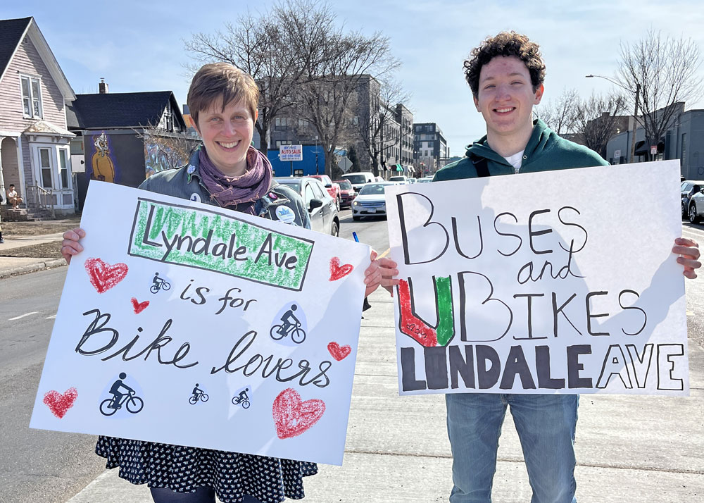 Two leaders from the Livable Lyndale campaign hold up signs at a March rally that say "Lyndale is for Bike Lovers" and "Buses and Bikes on Lyndale Ave"