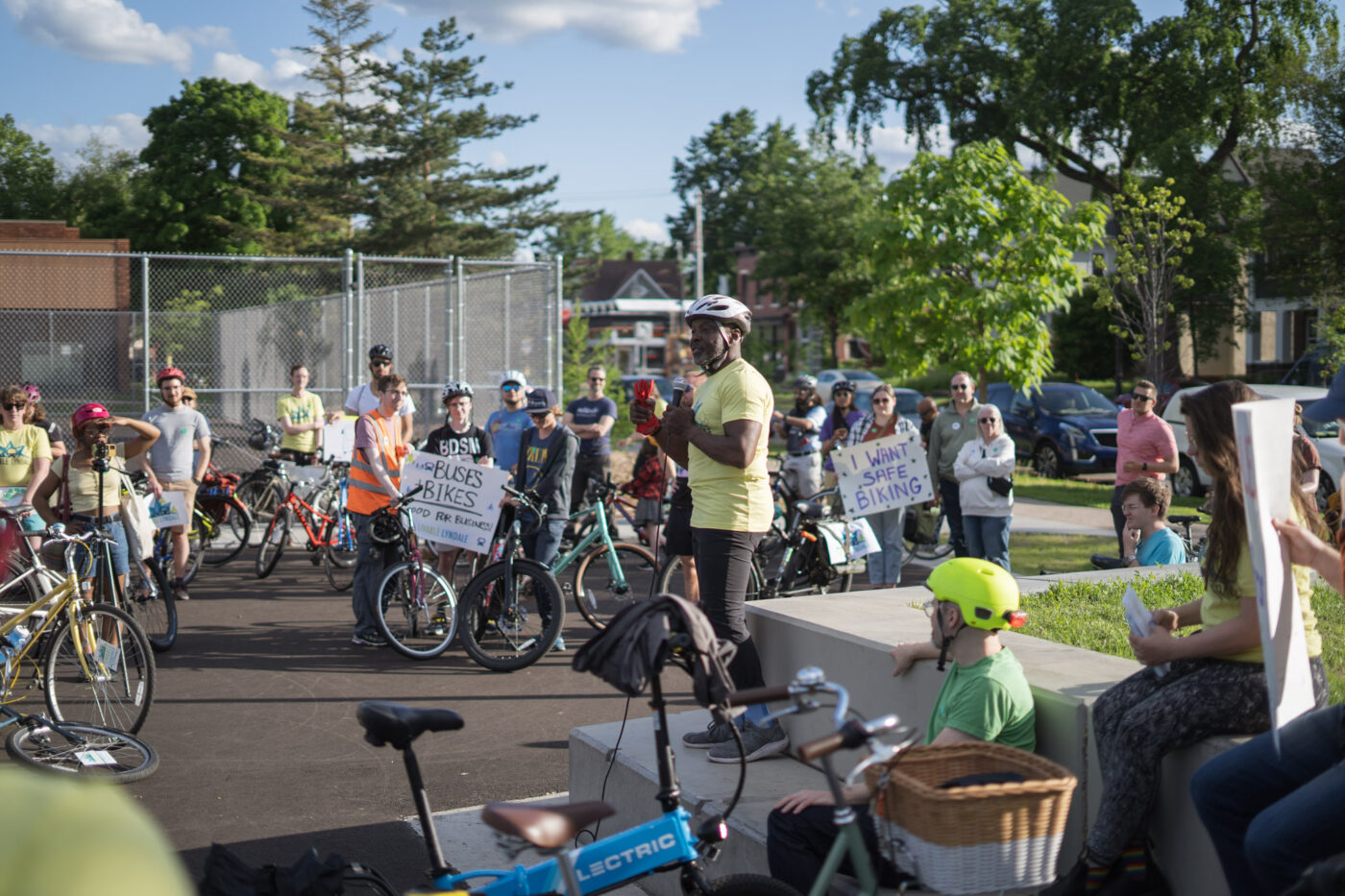 A Black man wearing a white bike helmet and bright green t-shirt speaks into a microphone inside a circle of people next to bicycles and holding signs for a Livable Lyndale