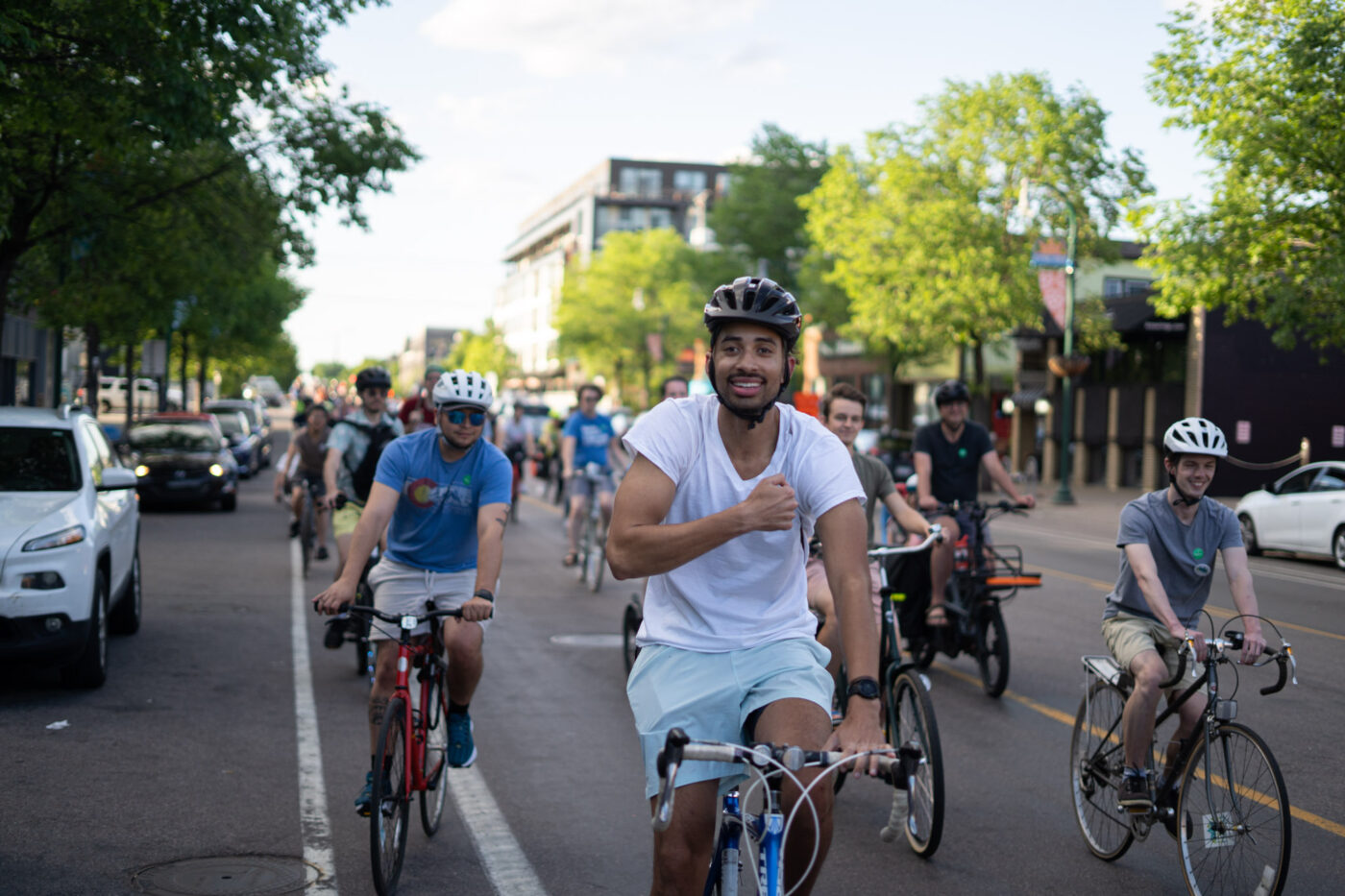 A young Black man wearing a white t-shirt and dark bike helmet at the front of a group of bicyclists with his fist to his heart in a gesture of affirmation