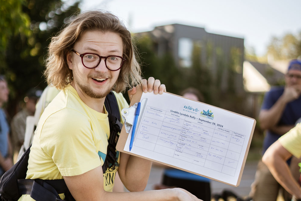 A male volunteer with chin length hair and red glasses poses playfully with the Livable Lyndale petition 