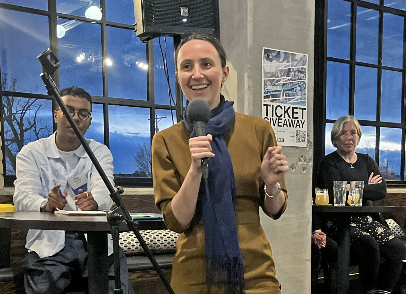 Rep. Katie Jones, a white woman wearing a brown dress and blue scarf, smiles as she addresses the crowd