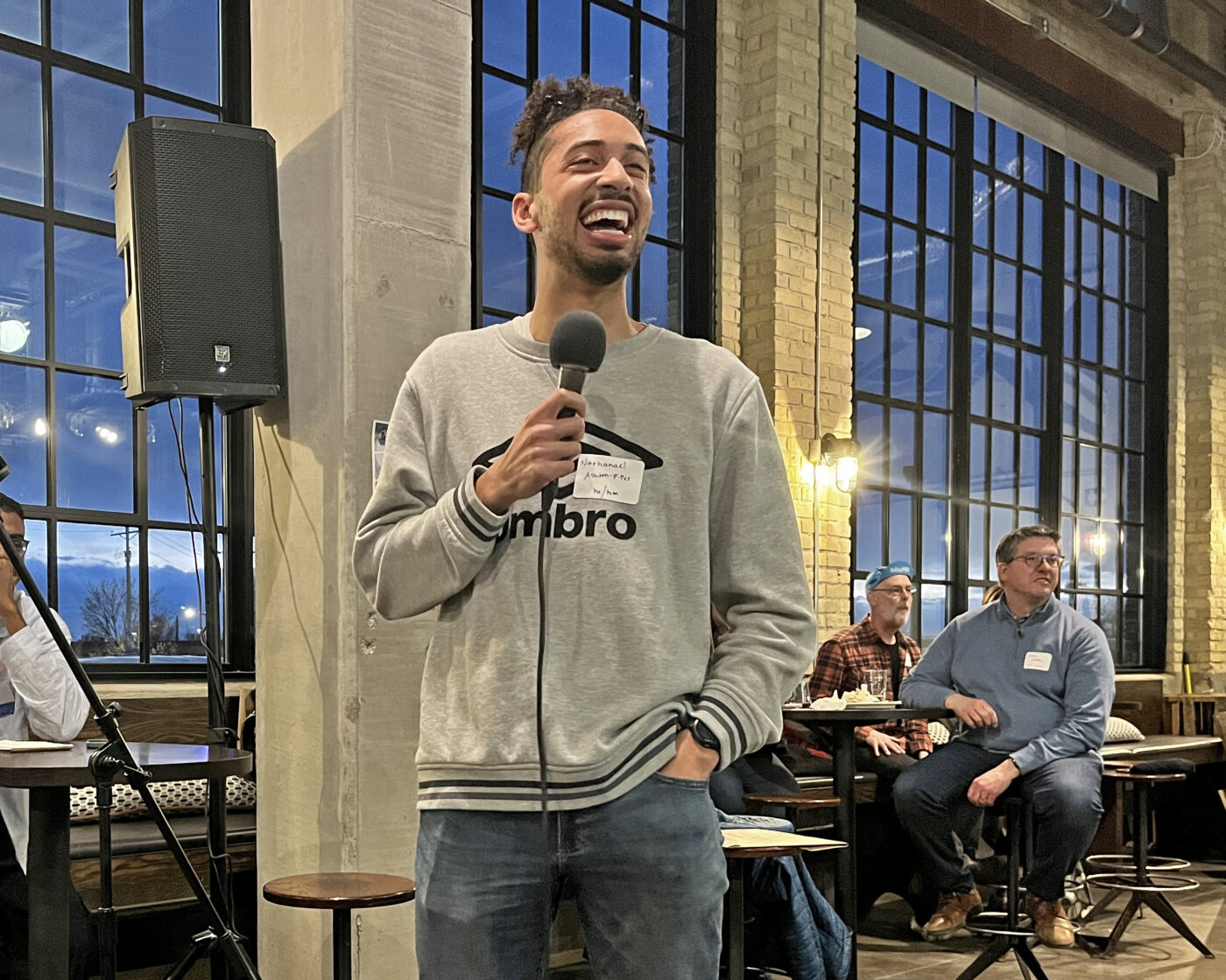 Nathanael Ashton-Piper, a tall. light-skinned Black man with facial hair, wearing a gray sweatshirt, laughs as he speaks to the crowd 